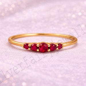 May include: A delicate gold ring featuring five oval-cut, vibrant red gemstones. The gemstones are set in a row across the top of the ring, creating a linear design. The ring's band is thin and polished, enhancing the focus on the gemstones.