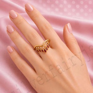 May include: A gold V-shaped ring featuring a series of dangling marquise-cut red stones and small round clear stones. The ring has a polished gold band that follows a chevron silhouette, with the stones suspended from the lower edge of the band.