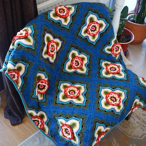May include: A crochet blanket featuring a repeating pattern of square motifs. Each square contains a central three-dimensional flower in shades of red, white, and yellow, surrounded by olive green and bright blue yarn in a geometric design.