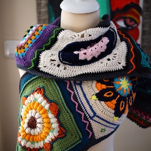 May include: A colorful, handmade crochet shawl. The shawl features a variety of geometric patterns and floral motifs in shades of green, orange, blue, purple, and white. The shawl is displayed on a mannequin, showcasing its intricate design and texture.