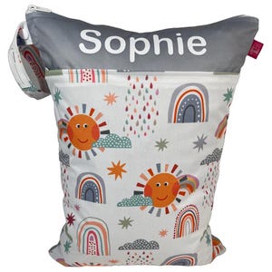Könnte beinhalten: Eine weiße Stofftasche mit einem bunten Muster aus Regenbogen, Sonnen, Wolken und Sternen. Die Tasche hat einen grauen oberen Teil mit dem Namen "Sophie" in Weiß. Eine kleinere, passende Tasche ist seitlich angebracht.
