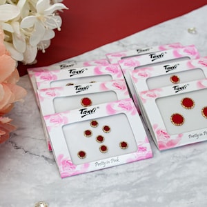 May include: A set of six red and gold bindi stickers, packaged in a clear plastic bag with a pink and white background. The packaging features the text "Tanvi" and "Pretty in Pink".