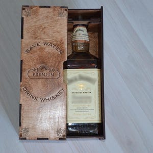 May include: A whiskey bottle presented in an open wooden box. The box is engraved with the words "SAVE WATER DRINK WHISKEY" and "PREMIUM". The bottle has a white label with black text. The box is open, revealing the bottle.