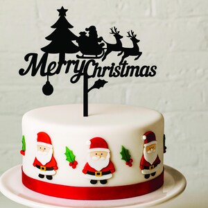 May include: A white cake decorated with Santa Claus figures and a red ribbon base. A black topper displays a Christmas tree, Santa in a sleigh, reindeer, and the words "Merry Christmas".