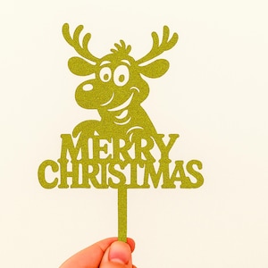 May include: A gold glitter Christmas cake topper featuring a smiling reindeer with antlers and the words "MERRY CHRISTMAS". The topper is held by a hand against a cream-colored background.