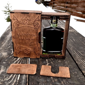 Op de afbeelding: Een houten doos met een fles donkere vloeistof erin. De doos heeft een scharnierend deksel en is gegraveerd met de woorden "Elegance Premium".