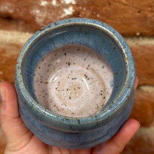 May include: A handmade ceramic bowl with a light blue exterior and a speckled, off-white interior. The bowl has a rounded shape with a textured, ridged design on the outside. The rim is a darker shade of blue.