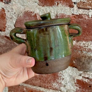 May include: A small, handmade ceramic pot with a lid. The pot is glazed in shades of green and brown, with two curved handles and a small knob on the lid. The base is a darker brown. The pot is held in a person's hand.