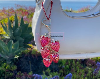 May include: A decorative bag charm featuring a cluster of six pink strawberry charms with gold-colored stems and a heart-shaped clasp. The charm is attached to a white handbag with a red beaded chain. The background includes greenery and a view of the ocean.