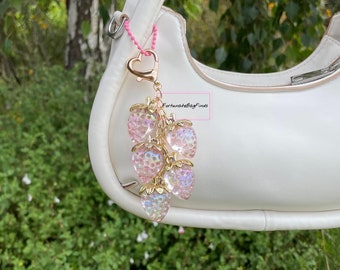 May include: A cluster of six pink strawberry charms with gold-colored stems and a heart-shaped clasp. The charms are attached to a pink beaded chain and hang from a white handbag. The background features green foliage.