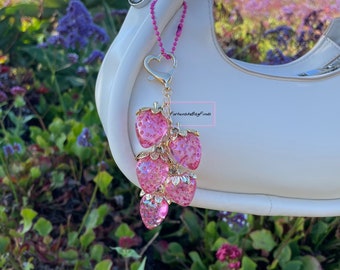 May include: A pink strawberry charm with gold accents hangs from a white handbag. The charm features six pink, glittery strawberries with gold stems and a heart-shaped clasp. The background shows a field of purple flowers and green foliage.