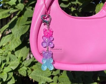 May include: A pink handbag with a charm attached to the handle. The charm is a stack of three gummy bear-shaped charms in shades of pink, purple, and blue.