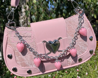 May include: Pink handbag with a crocodile texture, silver heart-shaped accents, and a chain adorned with strawberry charms. The bag has a flap closure and a shoulder strap. Silver heart-shaped studs are on the sides.