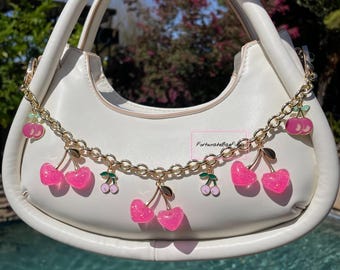 May include: A white handbag with a gold chain strap decorated with pink and green cherry charms. The charms are attached to the chain with gold rings.