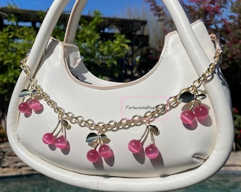 May include: White handbag with a gold chain strap and pink cherry charms. The chain has a small gold charm with the text "FortunateBags" on it.