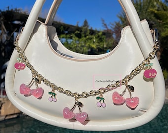 May include: Cream-colored handbag with a curved shape and a gold chain adorned with pink cherry charms. The bag has a top handle and a smooth texture. The background shows a blue sky and greenery.