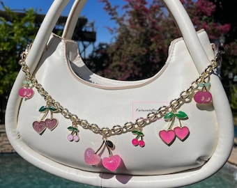 Könnte beinhalten: Eine weiße Handtasche mit einem goldenen Kettenriemen, der mit rosa und grünen Kirsch-Anhängern verziert ist. Die Anhänger sind mit Glitzer und herzförmigen Kirschen besetzt.