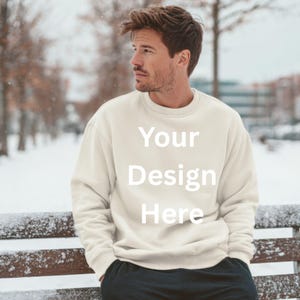 May include: Cream-colored sweatshirt with the text "Your Design Here" in white. The sweatshirt is worn by a person standing outside in a snowy environment. The person is wearing black pants and is leaning against a wooden bench.