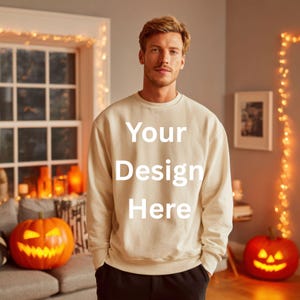 May include: Cream-colored sweatshirt with the text "Your Design Here" in white. The image features a person standing in front of a window and two carved pumpkins. The pumpkins are lit from within, creating a Halloween theme.