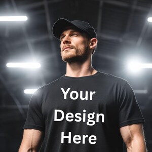 May include: Black t-shirt with the text "Your Design Here" in white. The person in the image is wearing a black baseball cap. The background is a blurred gym setting with bright lights.