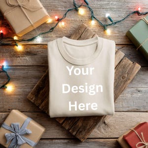 May include: A cream-colored sweatshirt with the text "Your Design Here" in white, resting on a rustic wooden board. The image is surrounded by wrapped gifts and a string of Christmas lights, creating a festive holiday theme.