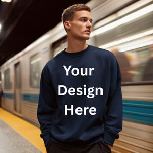 May include: A navy blue sweatshirt with the text "Your Design Here" in white. The sweatshirt is a crew neck style and is being worn by a person. The background shows a train and a yellow platform.