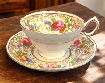 Puede incluir: Una taza de té y un platillo blancos con un patrón floral en tonos de rosa, amarillo y azul. El patrón incluye un borde dorado y detalles intrincados.