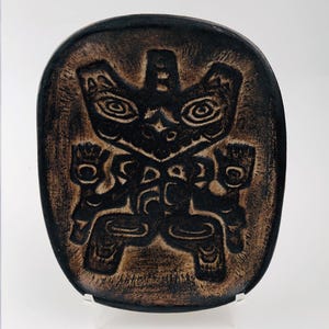May include: A dark brown, oval ceramic plate with a carved design. The design features a stylized figure with large eyes, hands, and feet. The plate has a textured surface and is displayed on a clear stand. The theme is Native American art.