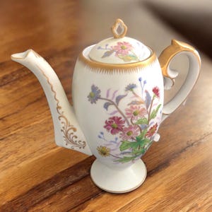 May include: A white porcelain coffee pot with a floral design featuring pink, purple, and yellow flowers. The pot has a gold handle, spout, and trim. The lid is topped with a gold finial. The pot is approximately 7 inches tall.