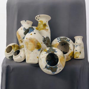 May include: A collection of seven ceramic vases in various sizes and shapes. The vases are decorated with a white glaze and splatters of blue, yellow, and brown paint.