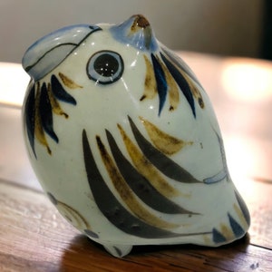 以下が含まれることがあります： 光沢のある白い釉薬が施された小さな陶器の鳥の置物。表面には、濃い青、茶色、黒の色合いで手描きされた抽象的な模様が描かれており、目立つ円形の目と様式化された翼の模様が特徴です。