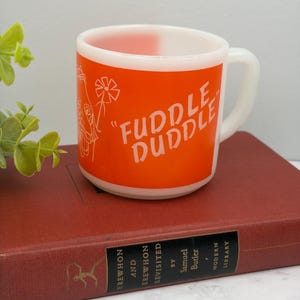 May include: A white milk glass mug featuring a bright orange rectangular panel with the text "FUDDLE DUDDLE" printed in white lettering. The design includes a stylized line drawing of a person holding a flower on the left side of the orange panel.