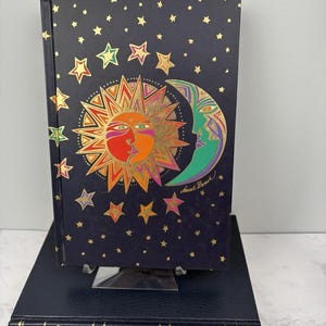 May include: A hardcover book with a dark blue cover featuring a colorful sun and moon design, surrounded by gold stars. The book is resting on a black book with gold lettering. The artist's signature is visible.