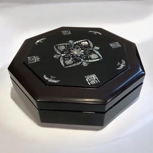 May include: An octagonal black lacquered box with a decorative floral design inlaid with mother-of-pearl. The box has a glossy finish and is likely used for jewelry or keepsakes. The lid features Asian characters.