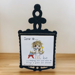 May include: A black cast iron hot pad with a white ceramic tile featuring a cartoon illustration of a woman on the phone. The tile reads "love is... keeping your cool when he brings a guest for dinner."