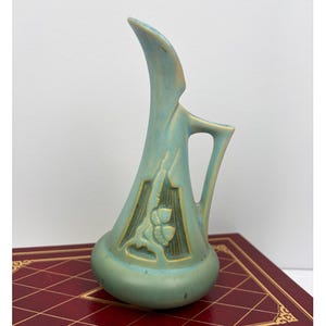 May include: A teal-colored art pottery vase with a slender, curved neck and a handle. The vase features a raised floral design and geometric patterns. The vase is approximately 10 inches tall.
