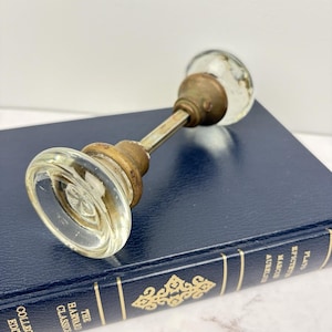 May include: A vintage glass doorknob with brass fittings lies on a dark blue book. The doorknob features clear, round glass handles and a matching base. The book is part of the Harvard Classics Collection.