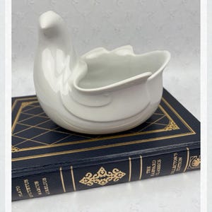May include: A white ceramic dove-shaped dish with a curved design. The dove is perched on a dark blue book with gold lettering. The dish has a smooth, glossy finish and a shallow bowl-like opening.