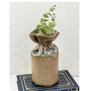 May include: A tan ceramic vase with a speckled texture and a gathered, pinched top rim. The vase features subtle blue and grey glaze accents near the neck and contains a small green artificial plant with rounded leaves extending upwards.