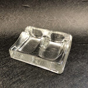 May include: A clear glass ashtray with two indented sections for cigarettes. The ashtray has a textured, grid-like pattern.