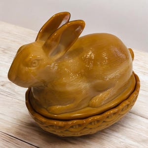 May include: A glazed ceramic bunny figurine in a warm brown hue. The bunny is seated in a basket-like base, with long ears and a detailed face. The figurine is likely a decorative item or a container, possibly for Easter.