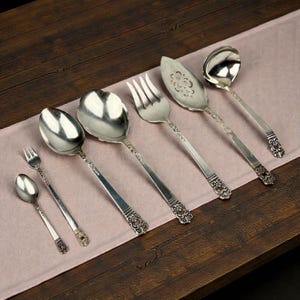 May include: A selection of antique silver serving utensils, including spoons, forks, and a pie server, displayed on a pink cloth. The silverware has ornate floral detailing on the handles and a polished surface, set against a dark wood background.