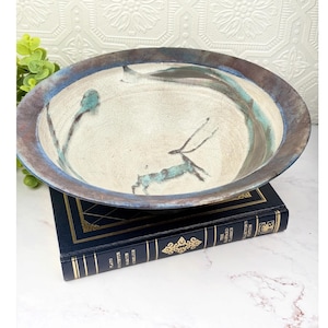 May include: A shallow ceramic bowl with a cream-coloured interior featuring a crackle glaze finish and a hand-painted teal deer illustration. The wide, dark brown rim has a metallic sheen, and the bowl rests on a dark blue hardcover book.