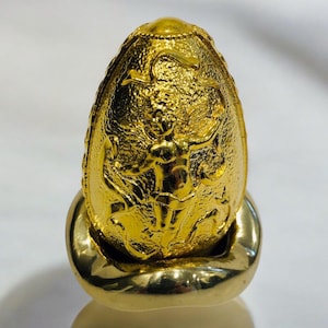 May include: A gold egg-shaped sculpture featuring a detailed relief of a figure with outstretched arms. The sculpture sits on a gold base, and the egg's surface has a textured, reflective finish. The design is ornate and decorative.