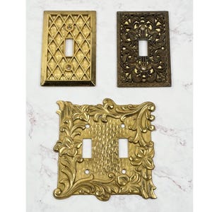 May include: Three metal light switch plates. The top left is a rectangular brass plate with a diamond grid pattern. The top right is a dark bronze rectangular plate with an intricate floral scrollwork design. The bottom is a large double switch plate.