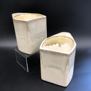 May include: Two off-white ceramic vases with a distinctive, angular shape. The vases have a glossy finish with a subtle, dripped glaze effect. The interior and top edges are a lighter shade, creating contrast. They sit on a clear acrylic stand.