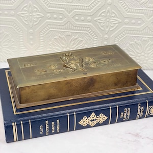 May include: A rectangular brass box with a hinged lid featuring an embossed relief of a seated figure surrounded by mushrooms. The metal surface has a weathered patina. The box rests on a dark blue hardcover book titled The Harvard Classics.