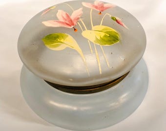 May include: A small, round, light blue trinket box with a frosted glass lid. The lid is decorated with hand-painted pink flowers and green leaves. The box has a brass-colored band around the middle.