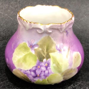 May include: A small, purple and white porcelain vase with a gold rim and a hand-painted design of purple violets and green leaves.