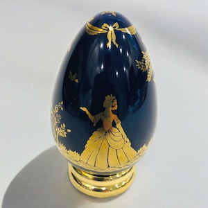 Puede incluir: Un huevo de cerámica azul oscuro con un soporte dorado. El huevo está decorado con una silueta dorada de una mujer con un vestido y otros diseños florales.